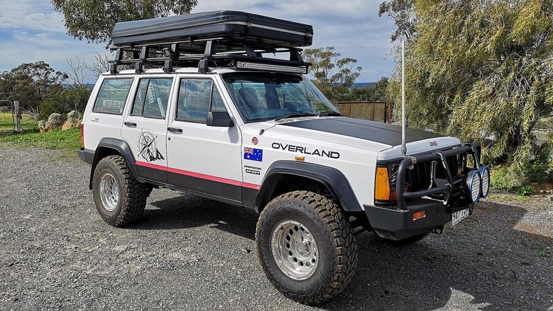 Modified white Jeep Cherokee XJ with a roof rack and steel bumper
